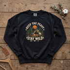Black Keep the Feast Stay Wild sweatshirt with a campfire, tent and pine trees, laid flat on a wooden table