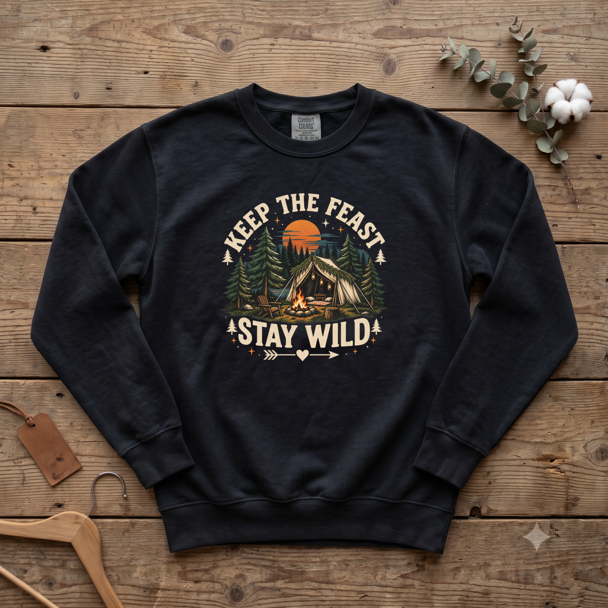Black Keep the Feast Stay Wild sweatshirt with a campfire, tent and pine trees, laid flat on a wooden table
