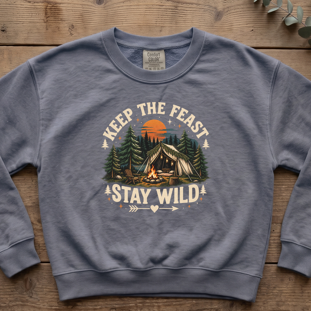 Blue jean Keep the Feast Stay Wild sweatshirt with a campfire, tent and pine trees, laid flat on a wooden table