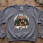 Blue jean Keep the Feast Stay Wild sweatshirt with a campfire, tent and pine trees, laid flat on a wooden table