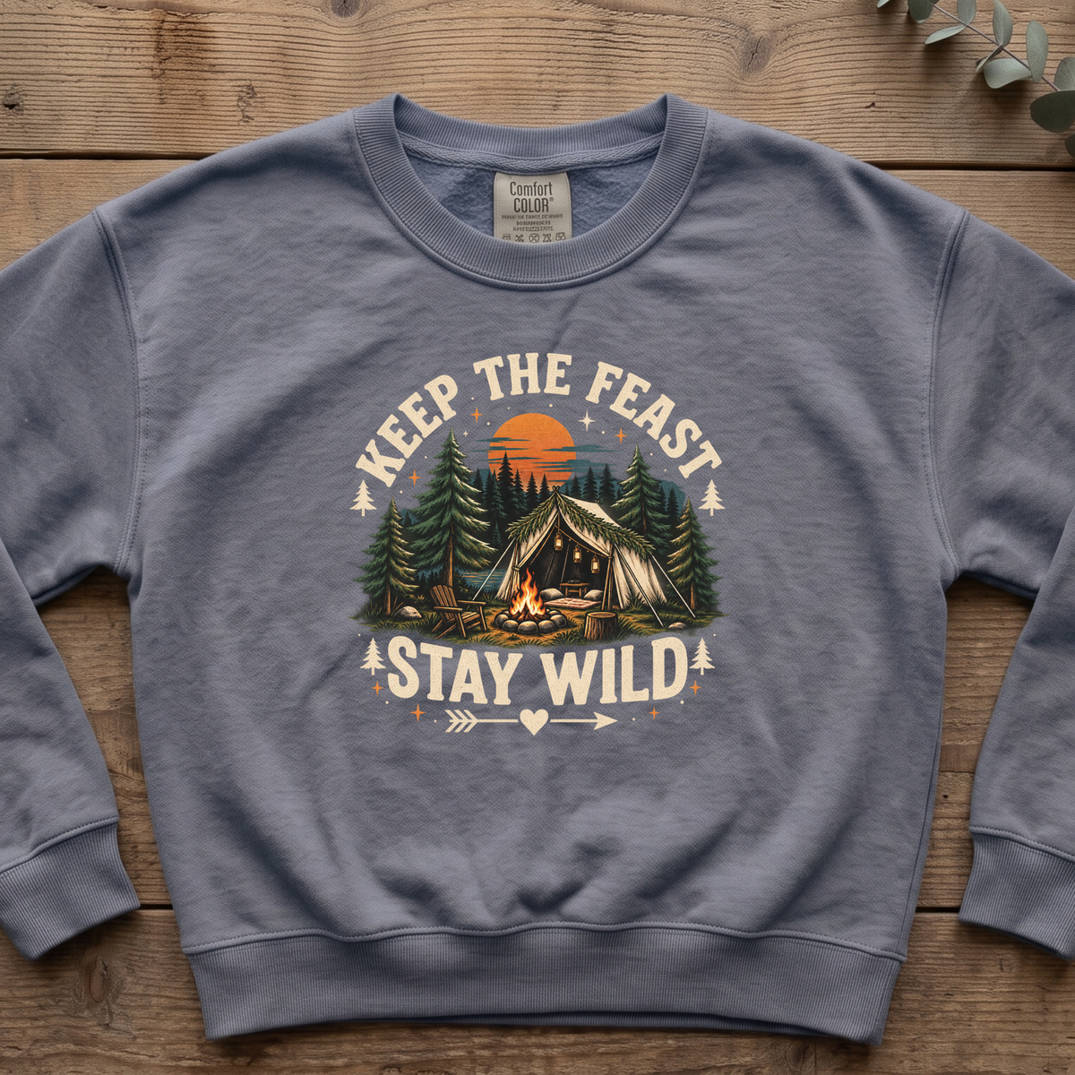 Blue jean Keep the Feast Stay Wild sweatshirt with a campfire, tent and pine trees, laid flat on a wooden table