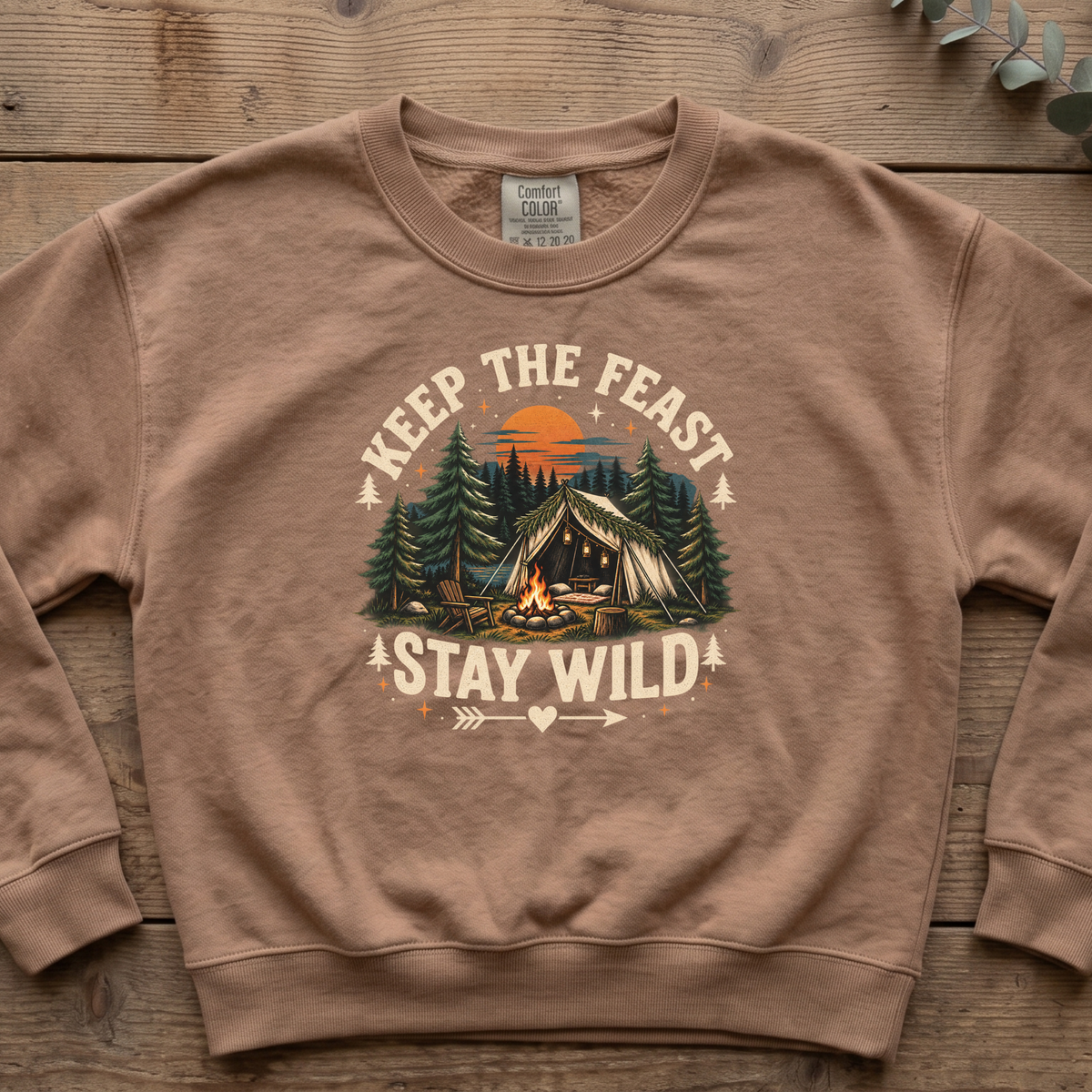 Espresso Keep the Feast Stay Wild sweatshirt with a campfire, tent and pine trees, laid flat on a wooden table