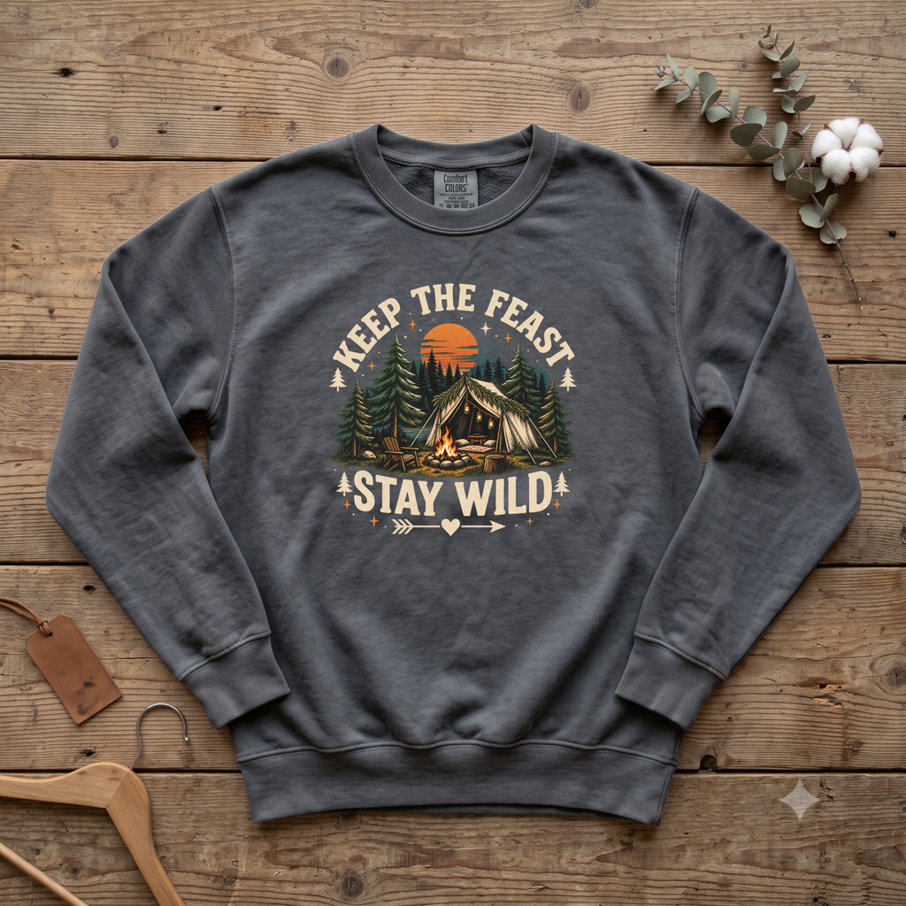 Pepper Keep the Feast Stay Wild sweatshirt with a campfire, tent and pine trees, laid flat on a wooden table