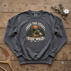 Pepper Keep the Feast Stay Wild sweatshirt with a campfire, tent and pine trees, laid flat on a wooden table