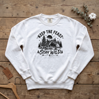 White Keep the Feast Stay Wild sweatshirt with a campfire, tent and pine trees, laid flat on a wooden table