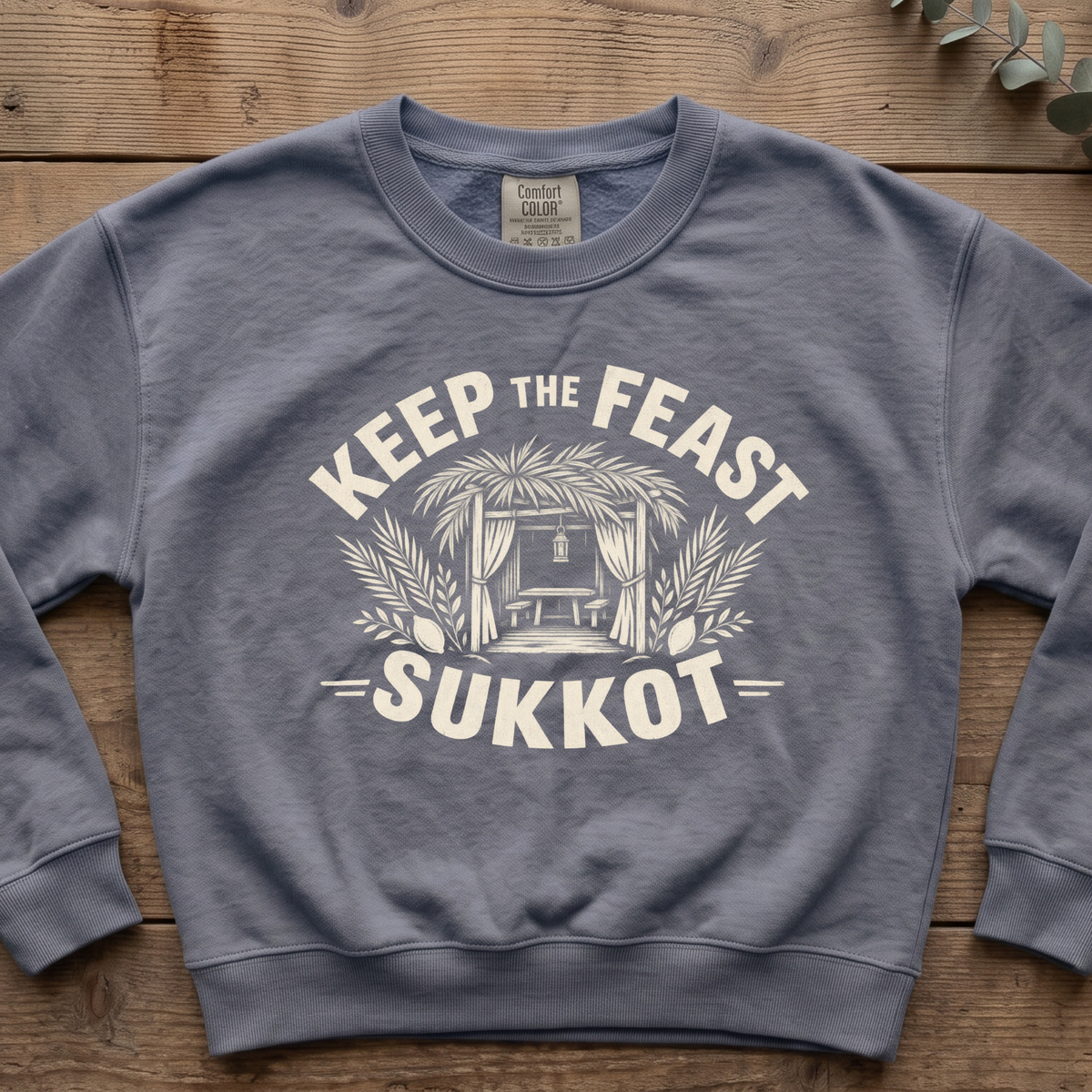 Blue jean Keep the Feast SUKKOT sweatshirt with a sukkah framed by palm branches, laid flat on a wooden table