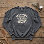 Pepper Keep the Feast SUKKOT sweatshirt with a sukkah framed by palm branches, laid flat on a wooden table