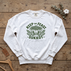White Keep the Feast SUKKOT sweatshirt with a sukkah framed by palm branches, laid flat on a wooden table