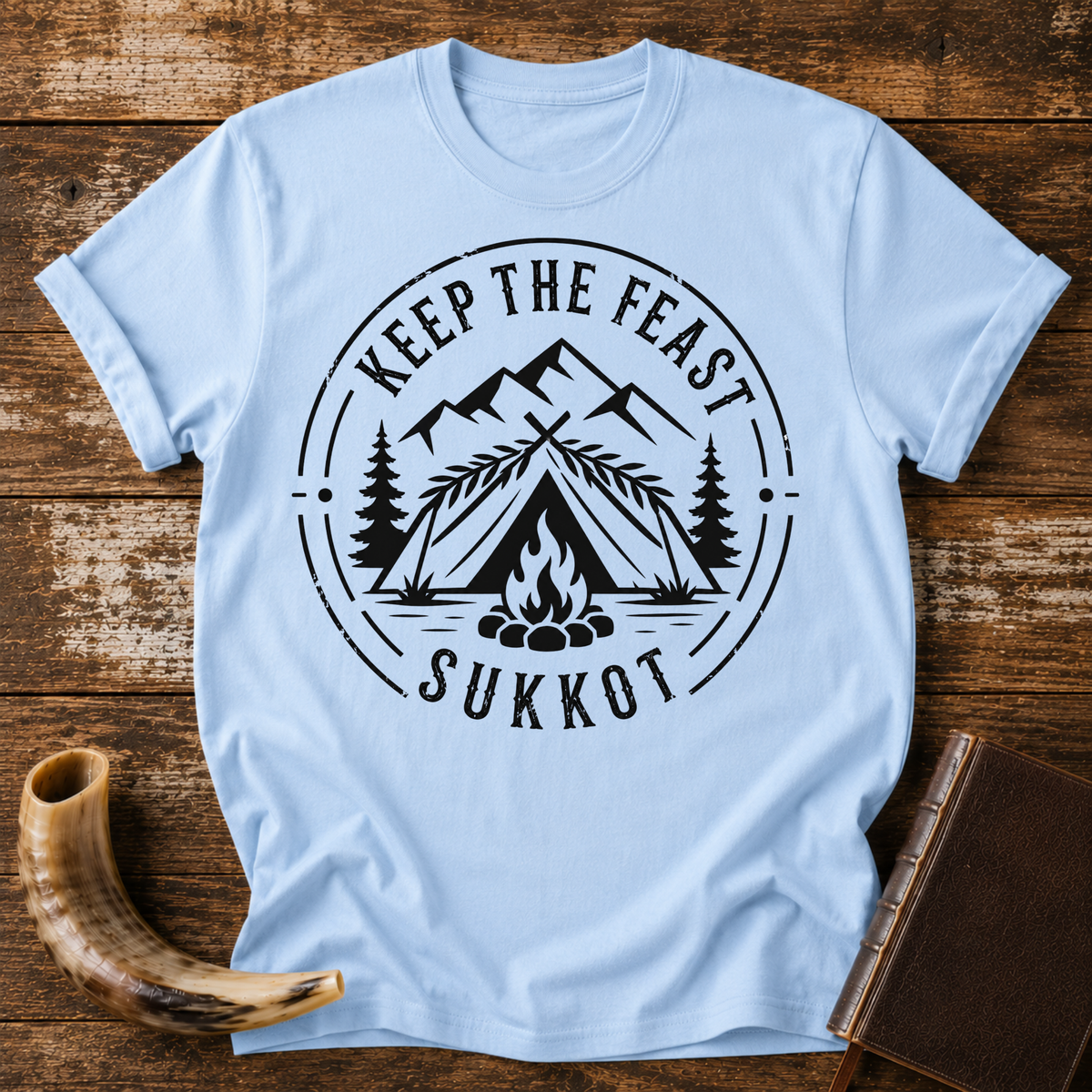 Light blue t-shirt with KEEP THE FEAST SUKKOT around a palm-roofed shelter, campfire and mountains, laid flat with a shofar and a leather book