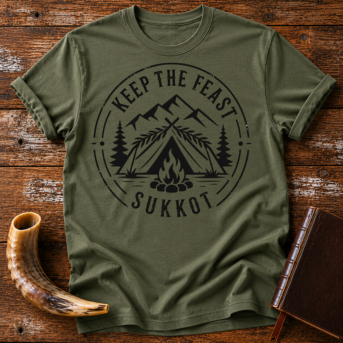 Military green t-shirt with KEEP THE FEAST SUKKOT around a palm-roofed shelter, campfire and mountains, laid flat with a shofar and a leather book