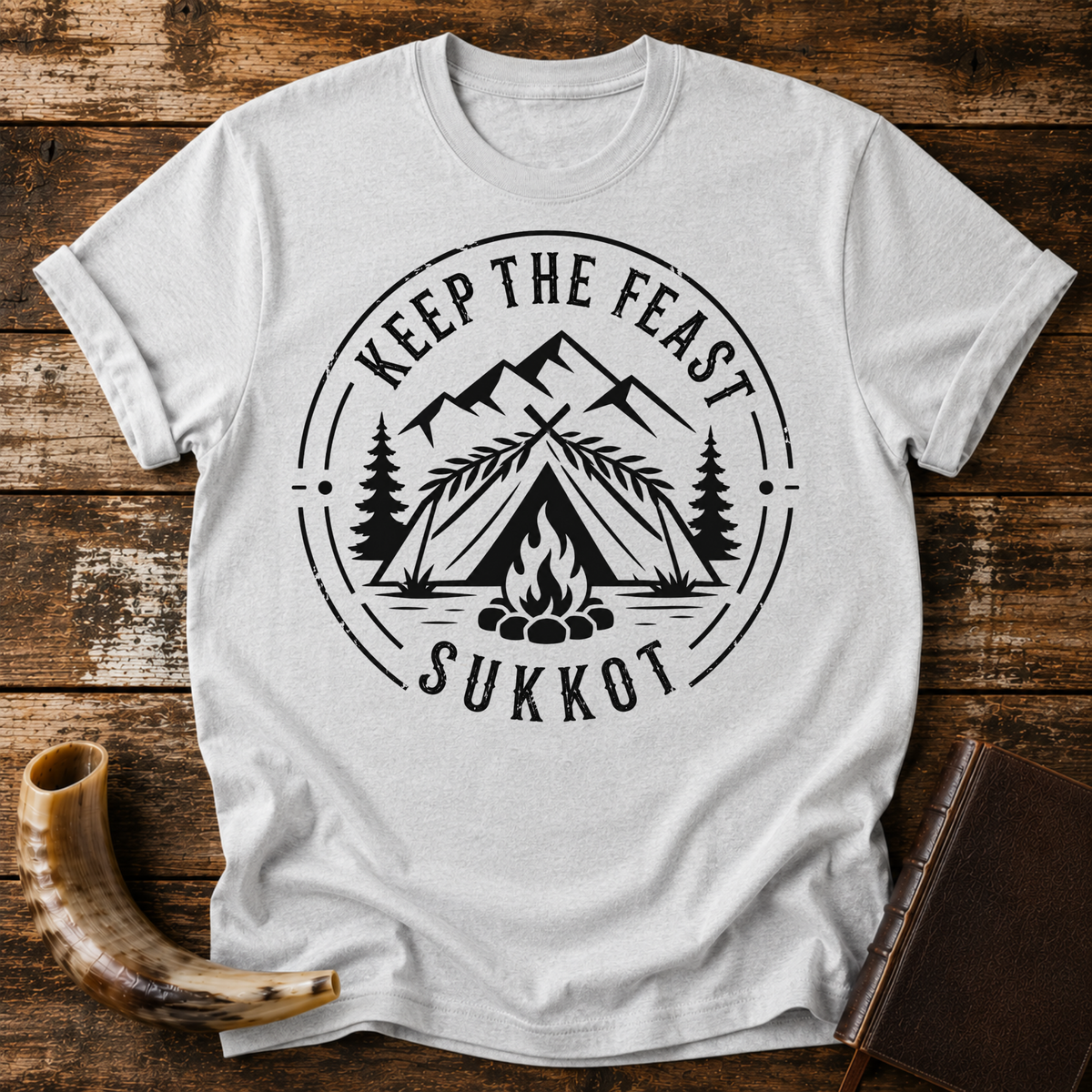 Sport grey t-shirt with KEEP THE FEAST SUKKOT around a palm-roofed shelter, campfire and mountains, laid flat with a shofar and a leather book