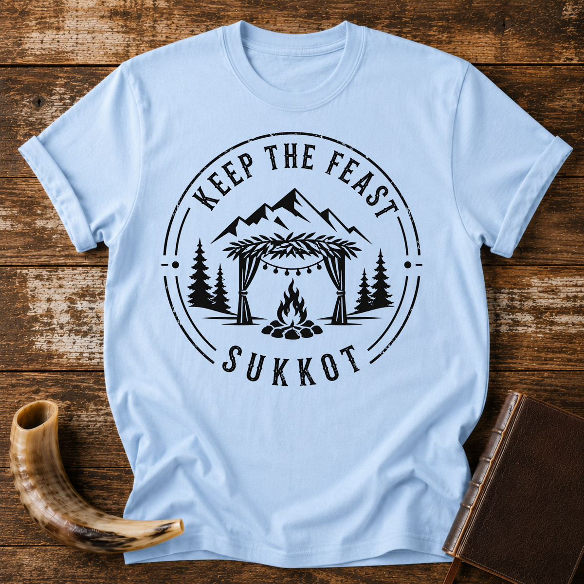 Light blue t-shirt with KEEP THE FEAST SUKKOT around a sukkah with string lights, campfire and pines, laid flat with a shofar and a leather book