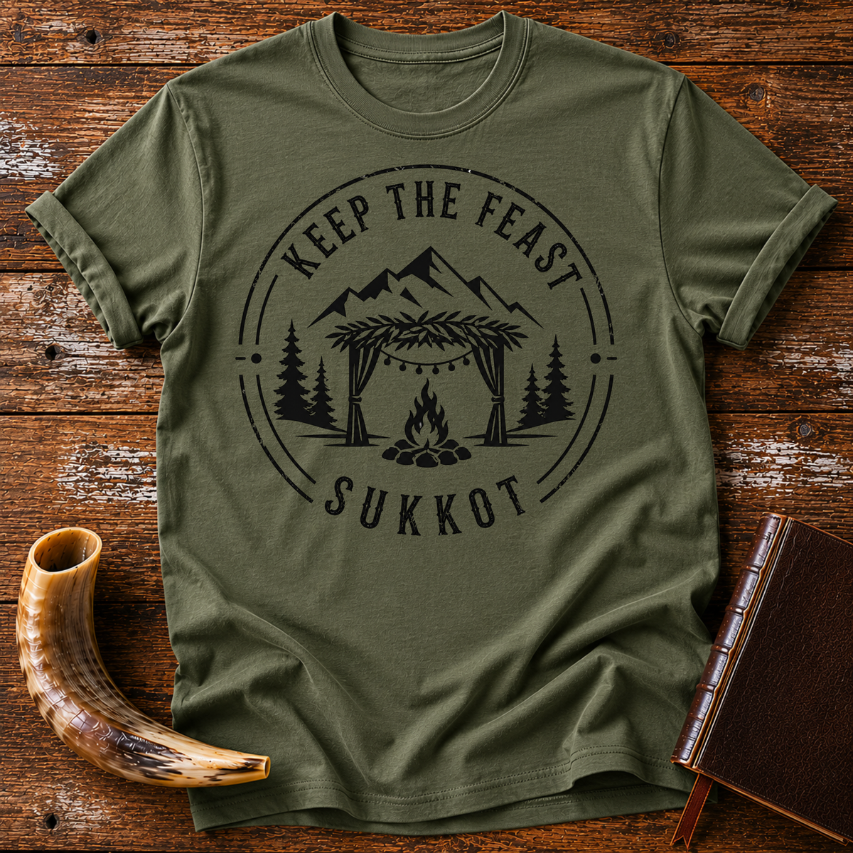 Military green t-shirt with KEEP THE FEAST SUKKOT around a sukkah with string lights, campfire and pines, laid flat with a shofar and a leather book