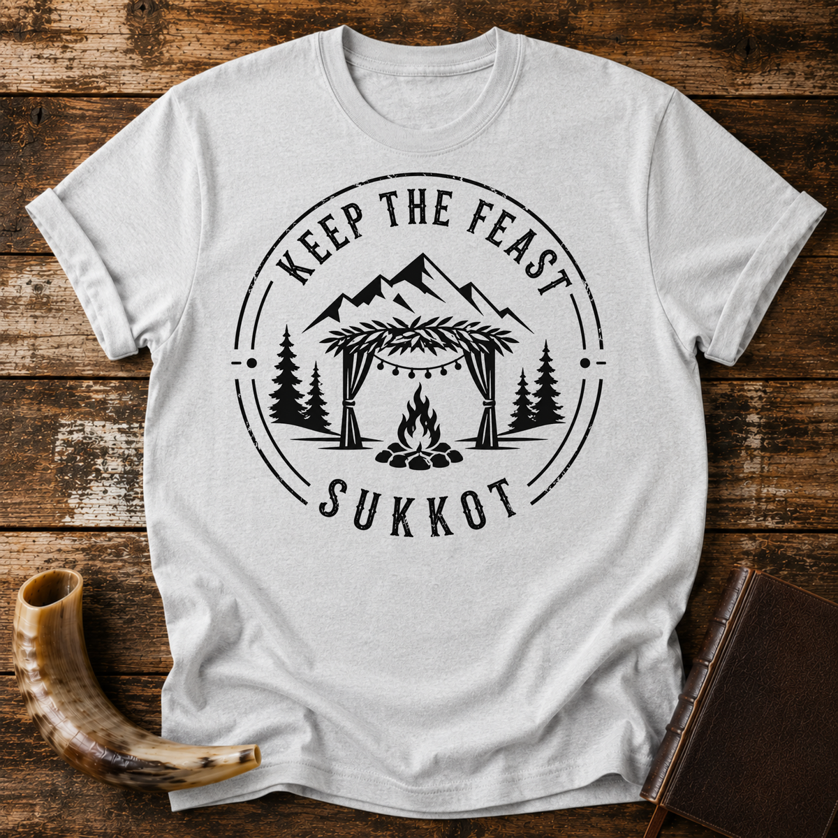 Sport grey t-shirt with KEEP THE FEAST SUKKOT around a sukkah with string lights, campfire and pines, laid flat with a shofar and a leather book