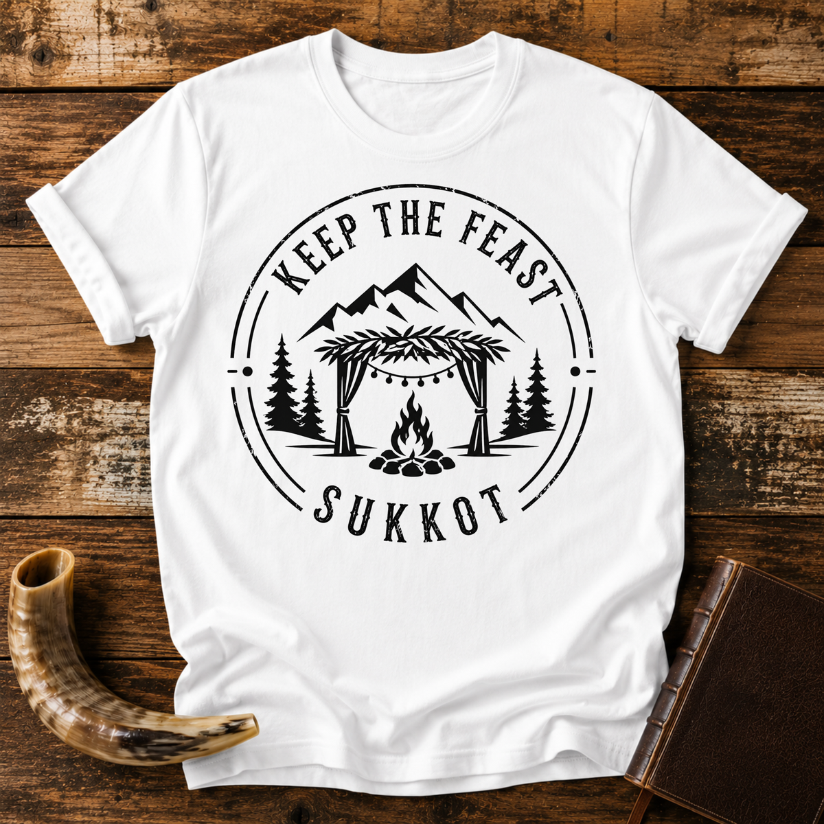 White t-shirt with KEEP THE FEAST SUKKOT around a sukkah with string lights, campfire and pines, laid flat with a shofar and a leather book
