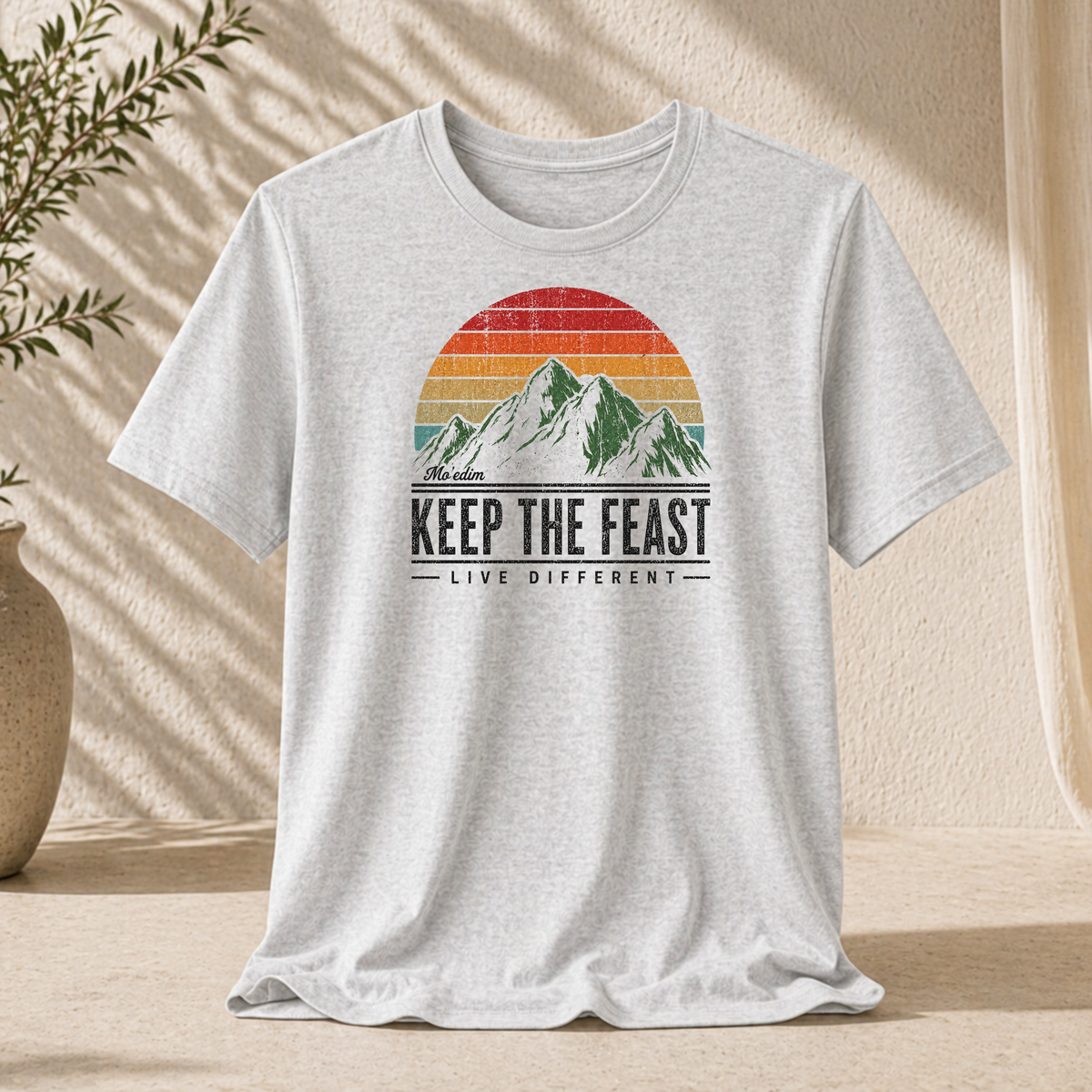 Ash gray Keep the Feast t-shirt with a retro sunset mountain graphic and "Live Different", hanging against a sunlit wall