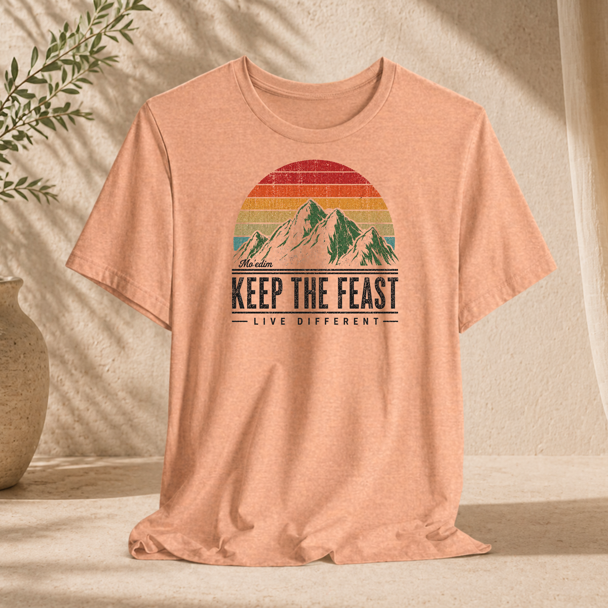 Heather peach Keep the Feast t-shirt with a retro sunset mountain graphic and "Live Different", hanging against a sunlit wall