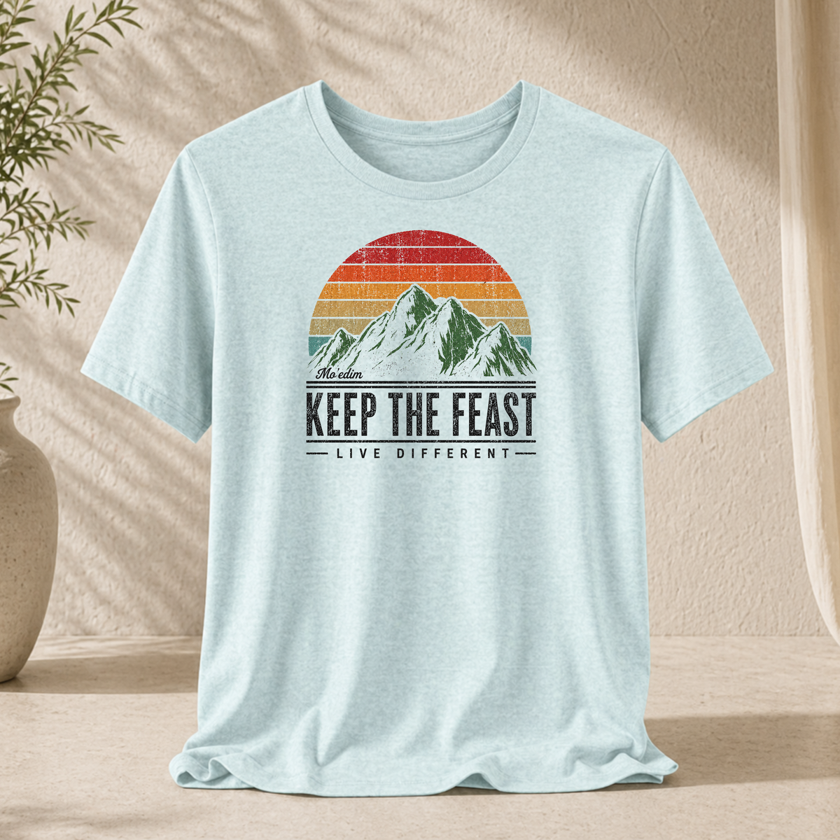 Heather ice blue Keep the Feast t-shirt with a retro sunset mountain graphic and "Live Different", hanging against a sunlit wall