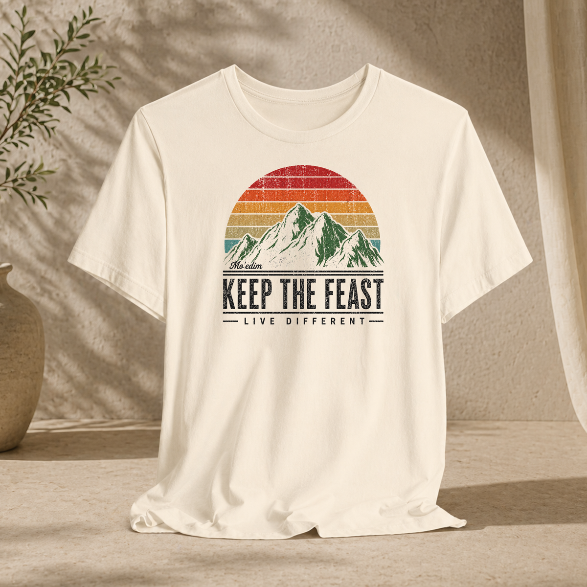 Natural cream Keep the Feast t-shirt with a retro sunset mountain graphic and "Live Different", hanging against a sunlit wall
