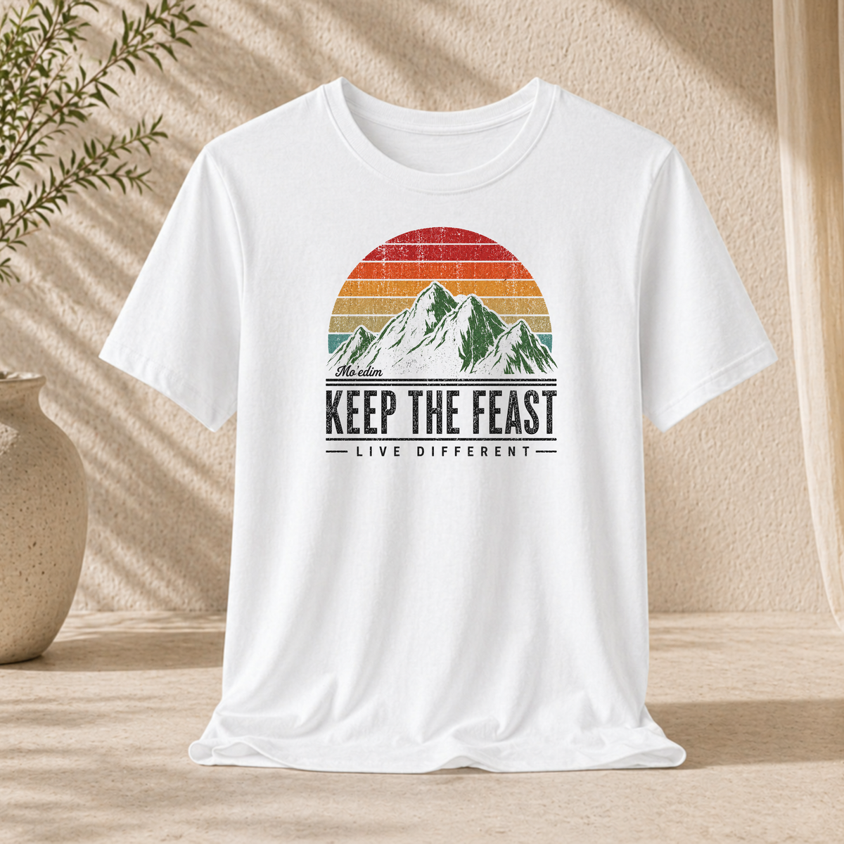 White Keep the Feast t-shirt with a retro sunset mountain graphic and "Live Different", hanging against a sunlit wall