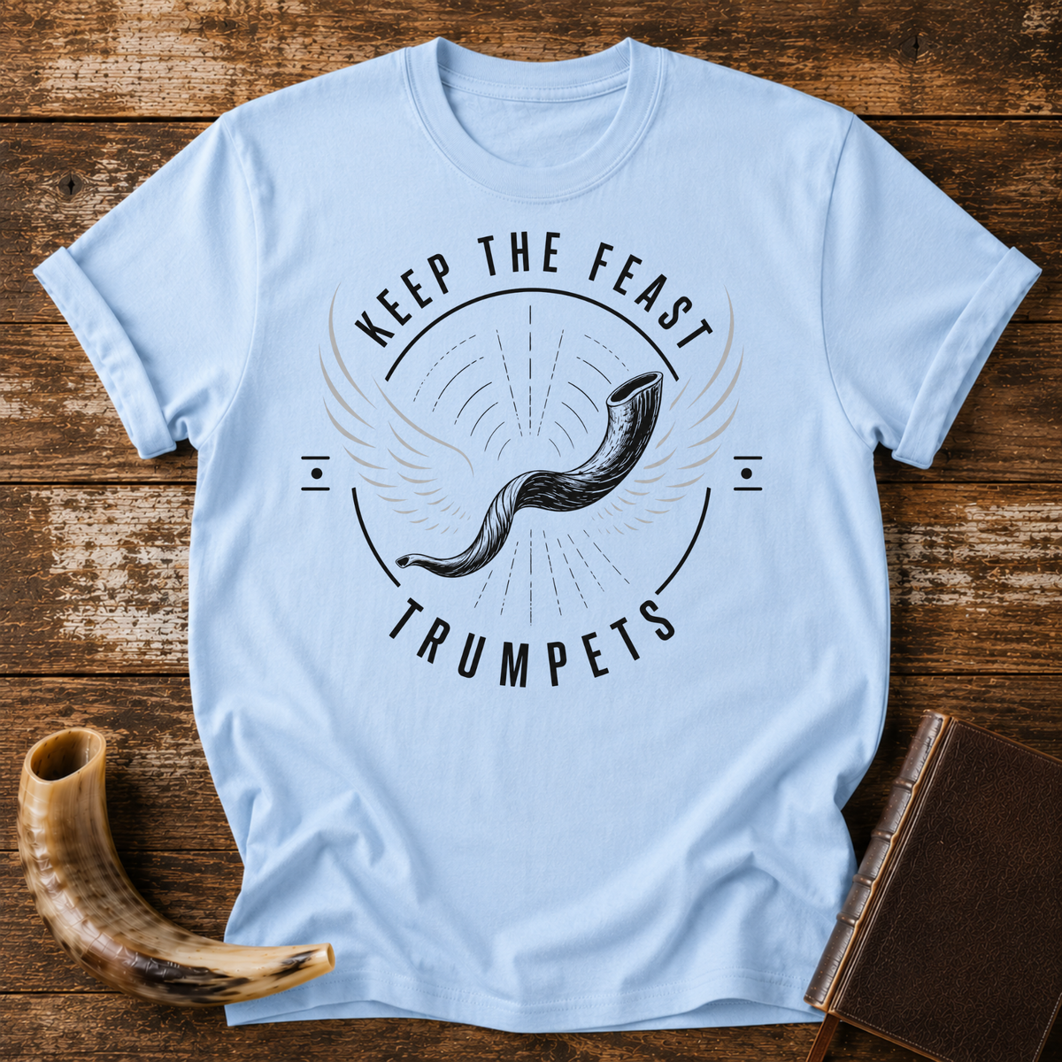 Light blue t-shirt with a winged shofar inside KEEP THE FEAST TRUMPETS, laid flat with a shofar and a leather book