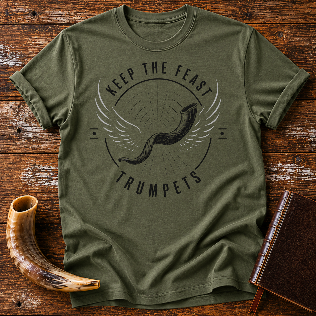 Military green t-shirt with a winged shofar inside KEEP THE FEAST TRUMPETS, laid flat with a shofar and a leather book