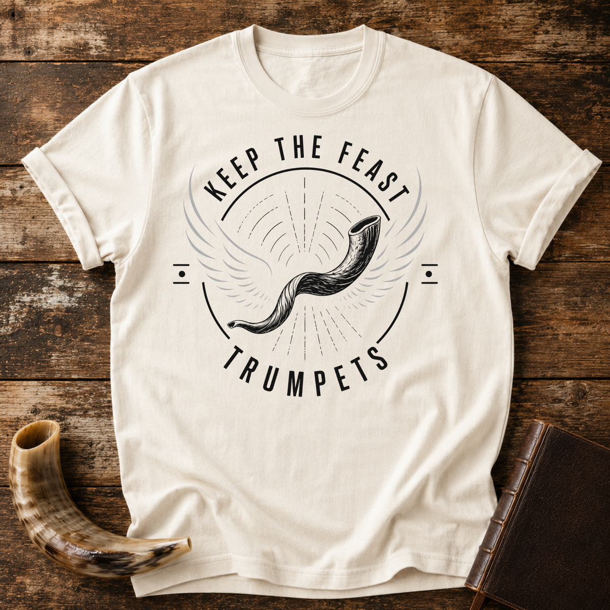 Natural cream t-shirt with a winged shofar inside KEEP THE FEAST TRUMPETS, laid flat with a shofar and a leather book