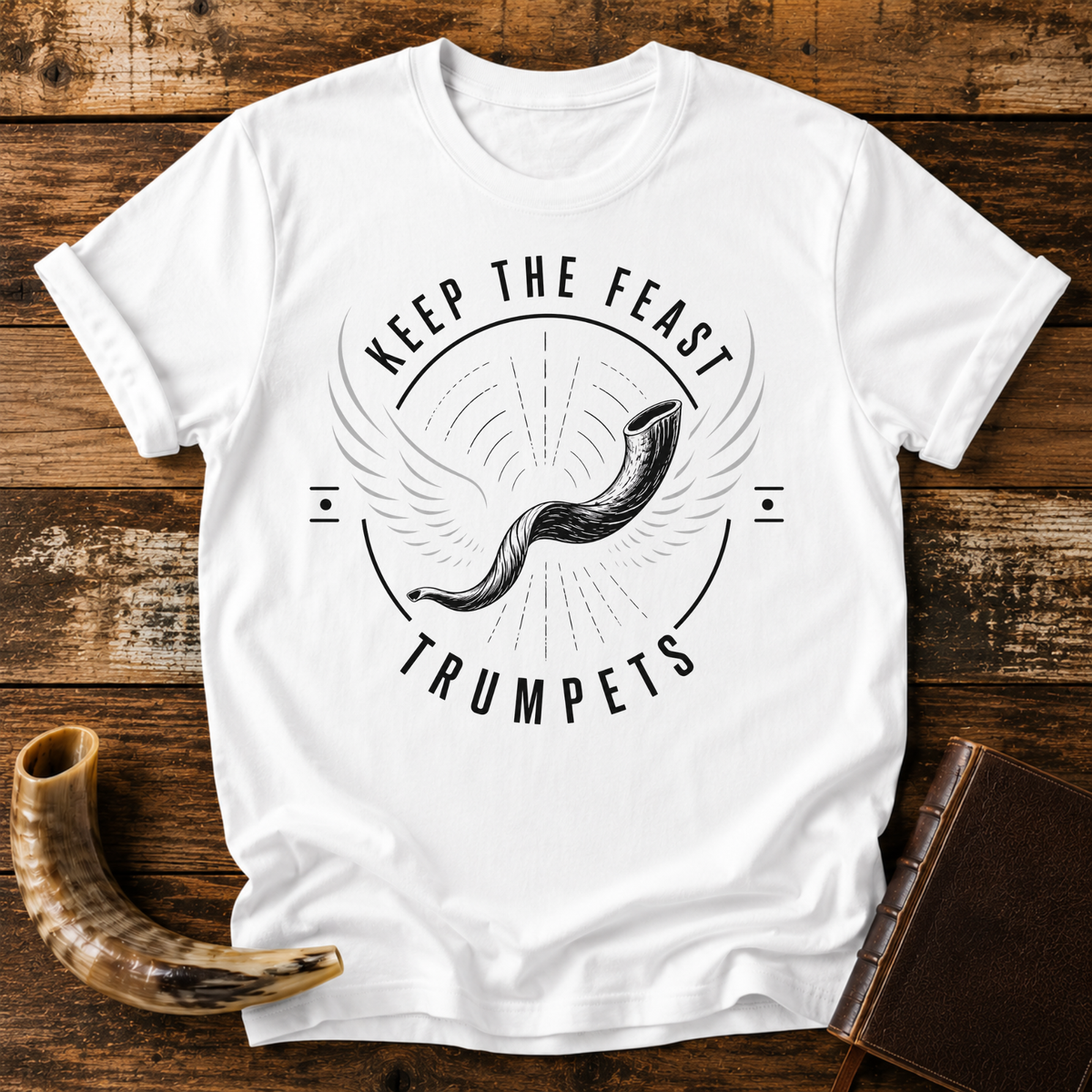 White t-shirt with a winged shofar inside KEEP THE FEAST TRUMPETS, laid flat with a shofar and a leather book