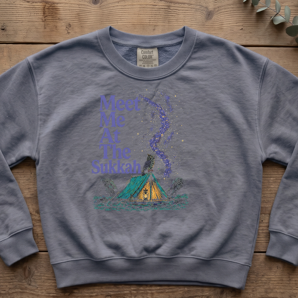 Blue jean Meet Me At The Sukkah sweatshirt with a purple starry sky over a glowing tent, laid flat on a wooden table