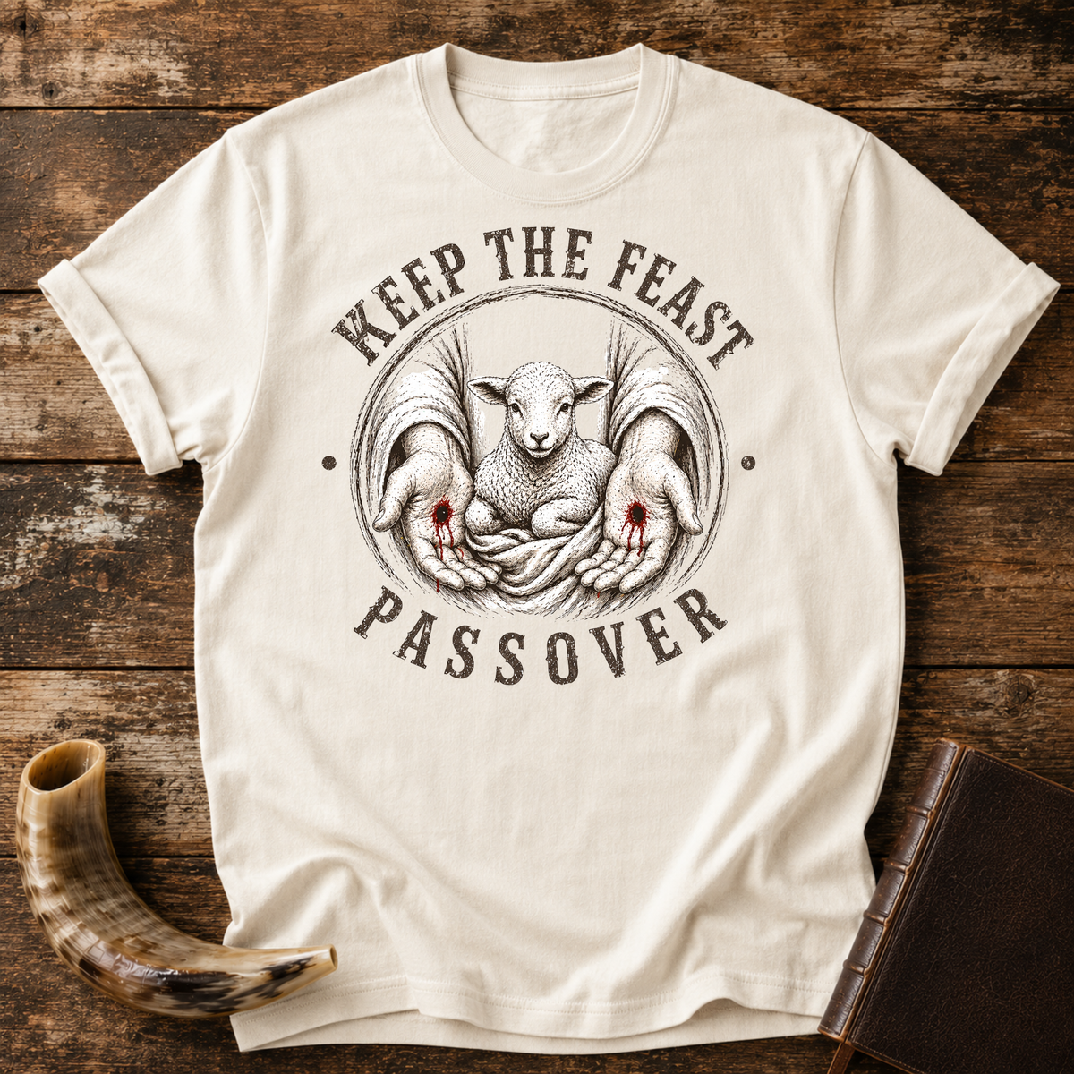 Natural cream t-shirt with KEEP THE FEAST PASSOVER around a lamb held in nail-scarred hands, laid flat with a shofar and a leather book