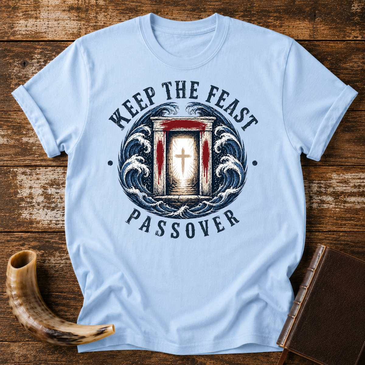 Light blue t-shirt with KEEP THE FEAST PASSOVER, a red-marked doorway with a cross and waves, laid flat with a shofar and a leather book