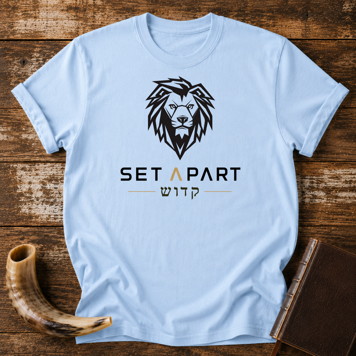 Light blue t-shirt with a large black lion head above SET APART and Hebrew lettering, laid flat with a shofar and a leather book