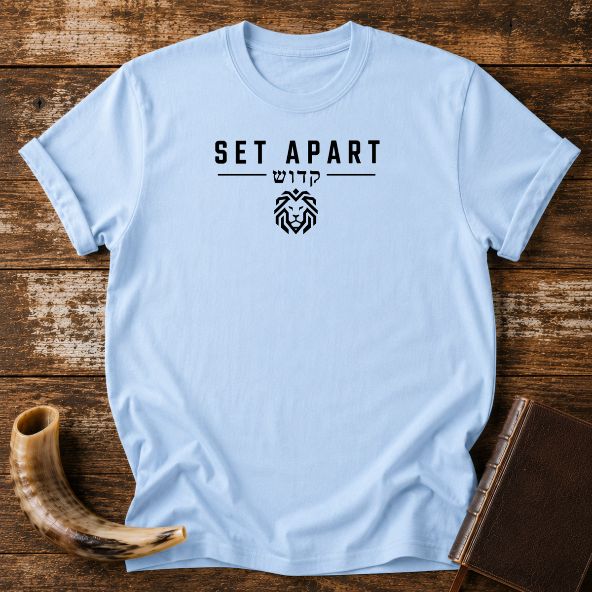 Light blue t-shirt with SET APART, Hebrew lettering and a small geometric lion head, laid flat with a shofar and a leather book