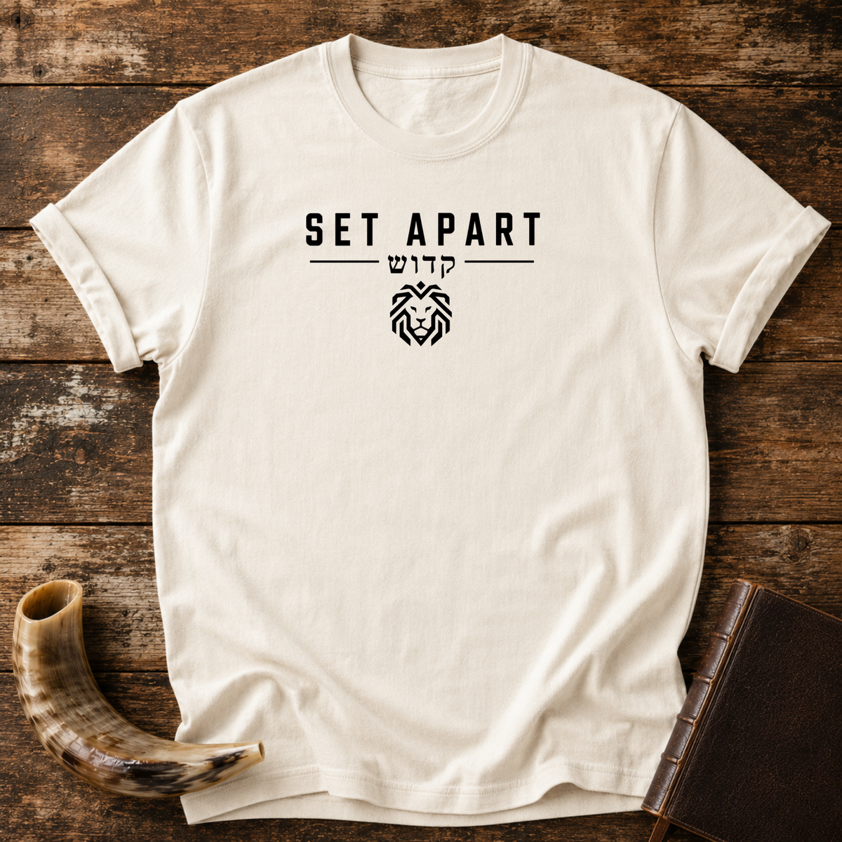 Natural cream t-shirt with SET APART, Hebrew lettering and a small geometric lion head, laid flat with a shofar and a leather book
