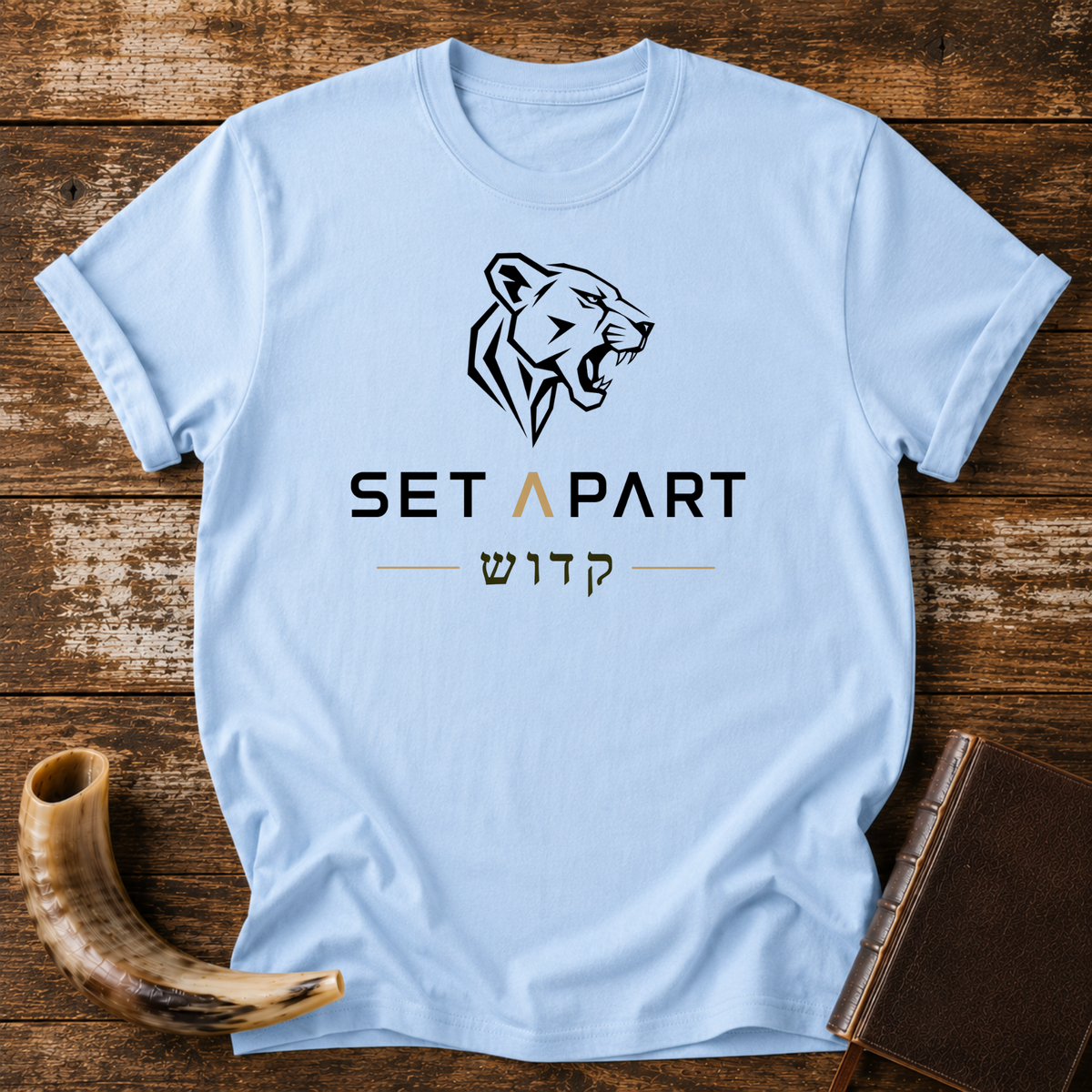 Light blue t-shirt with a roaring lioness head above SET APART and Hebrew lettering, laid flat with a shofar and a leather book