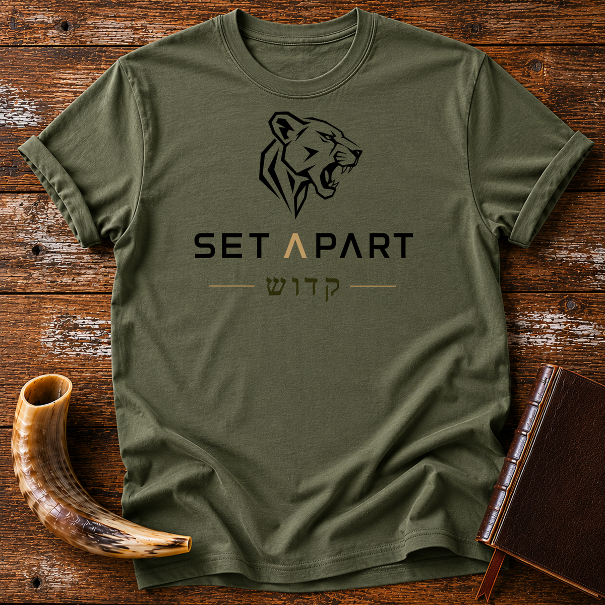 Military green t-shirt with a roaring lioness head above SET APART and Hebrew lettering, laid flat with a shofar and a leather book