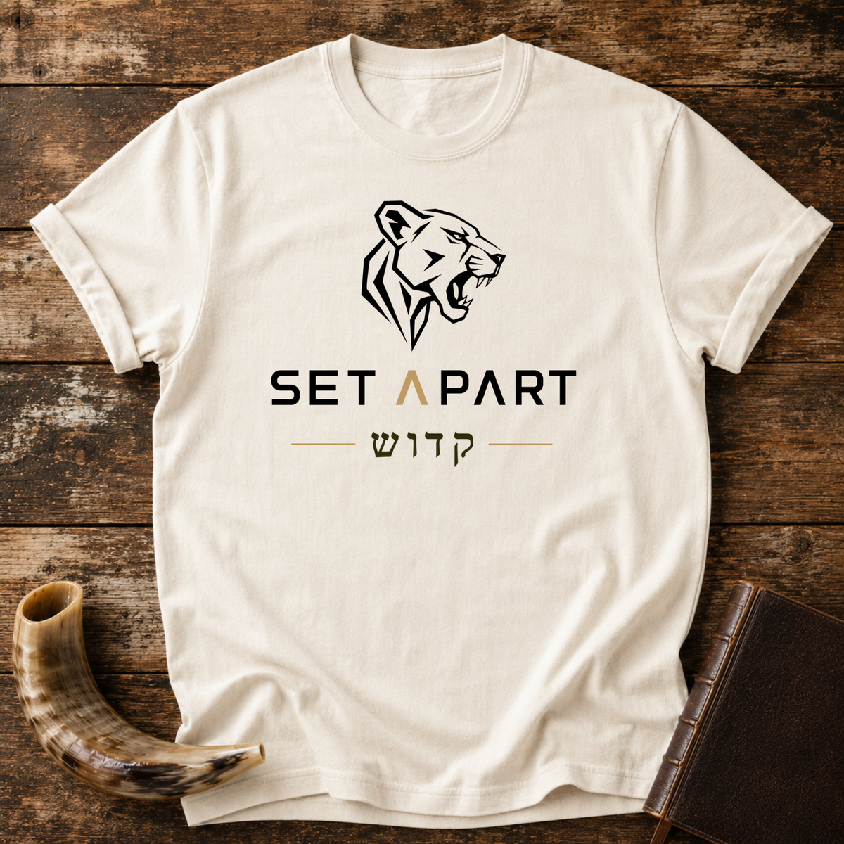 Natural cream t-shirt with a roaring lioness head above SET APART and Hebrew lettering, laid flat with a shofar and a leather book