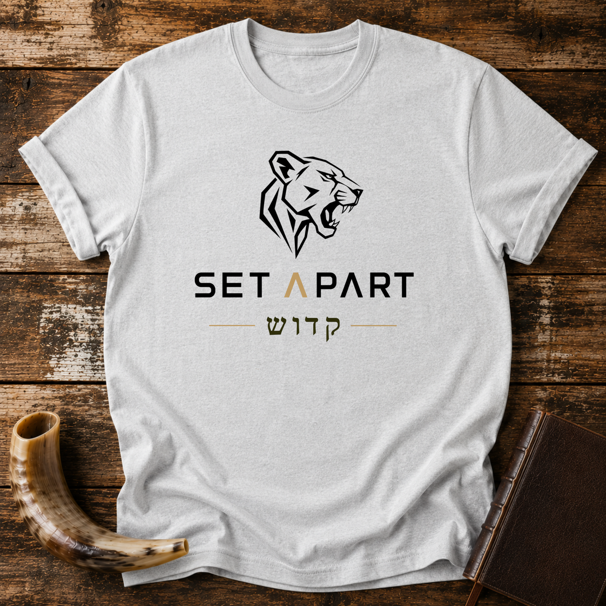 Sport grey t-shirt with a roaring lioness head above SET APART and Hebrew lettering, laid flat with a shofar and a leather book