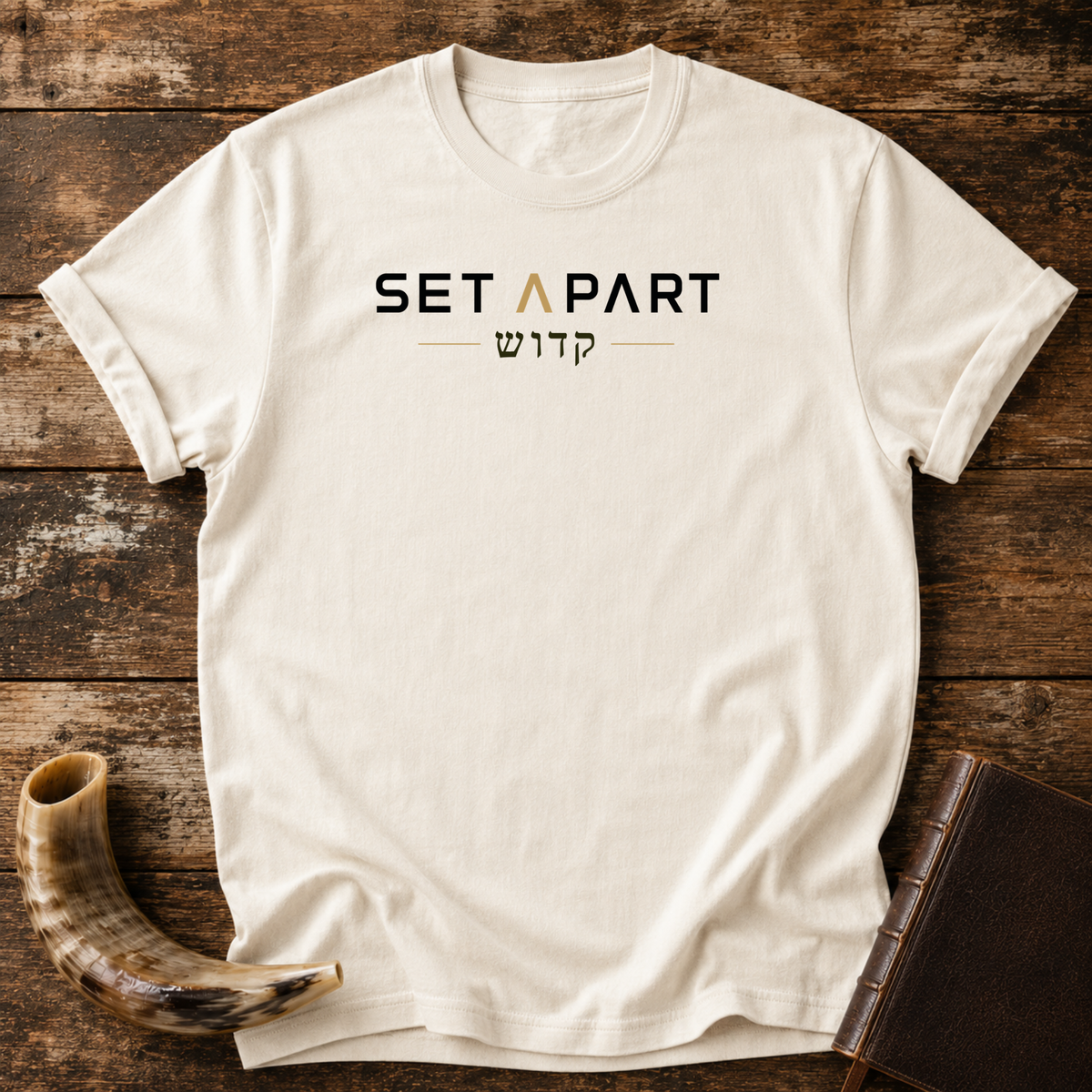 Natural cream t-shirt with SET APART text in black and gold over a line of Hebrew lettering, laid flat with a shofar and a leather book