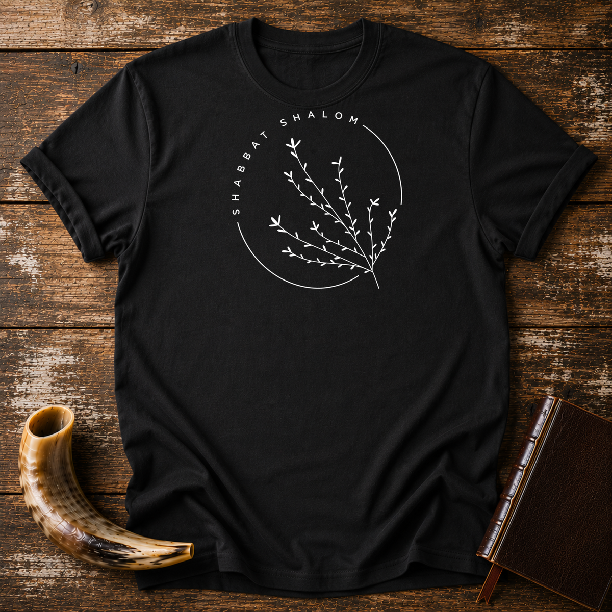 Black t-shirt with a thin SHABBAT SHALOM circle around a delicate branch, laid flat with a shofar and a leather book