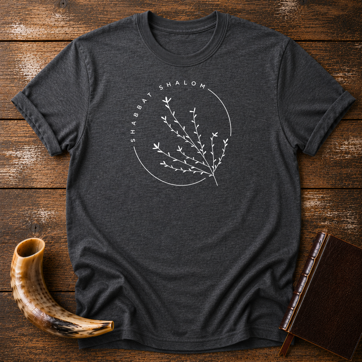 Dark heather gray t-shirt with a thin SHABBAT SHALOM circle around a delicate branch, laid flat with a shofar and a leather book