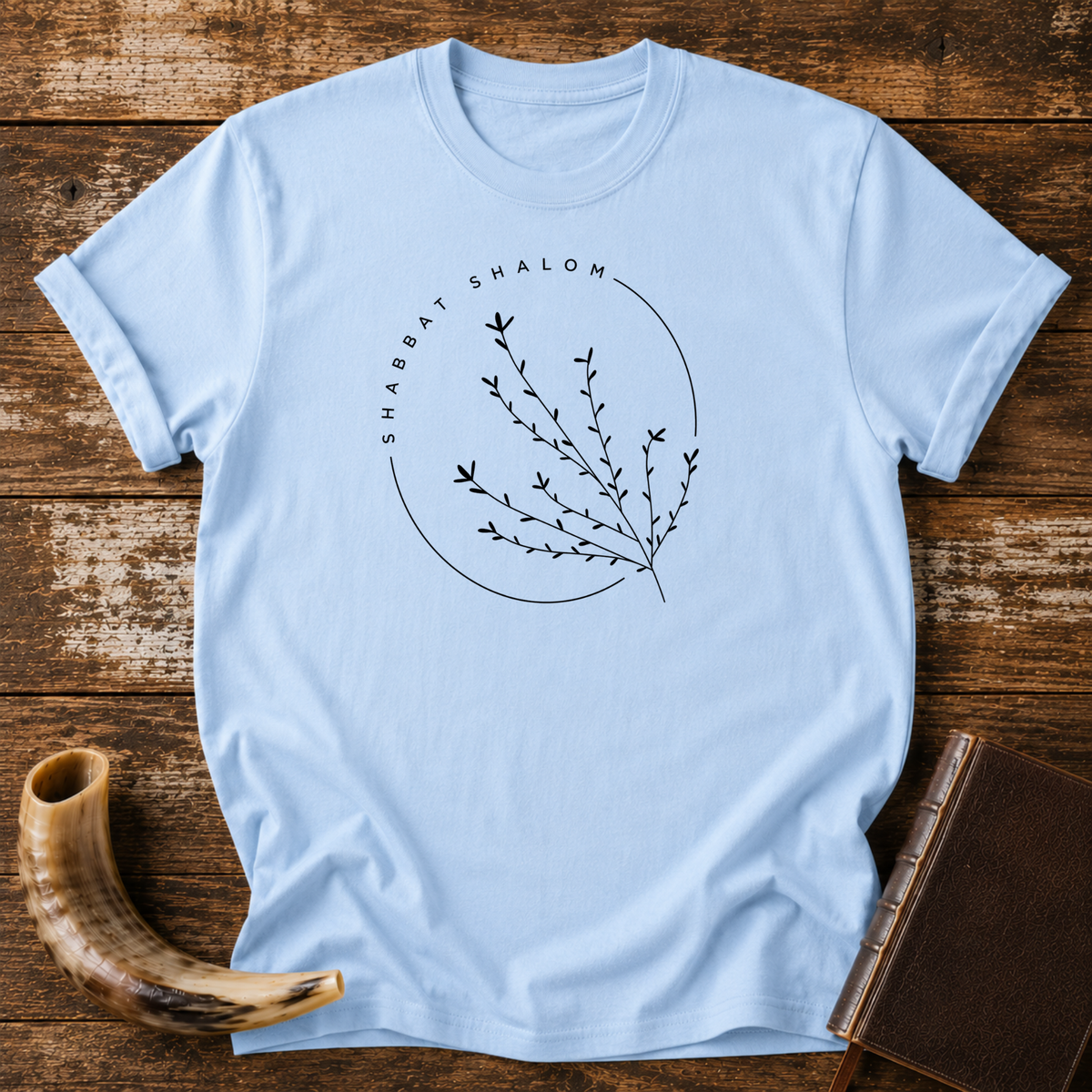 Light blue t-shirt with a thin SHABBAT SHALOM circle around a delicate branch, laid flat with a shofar and a leather book