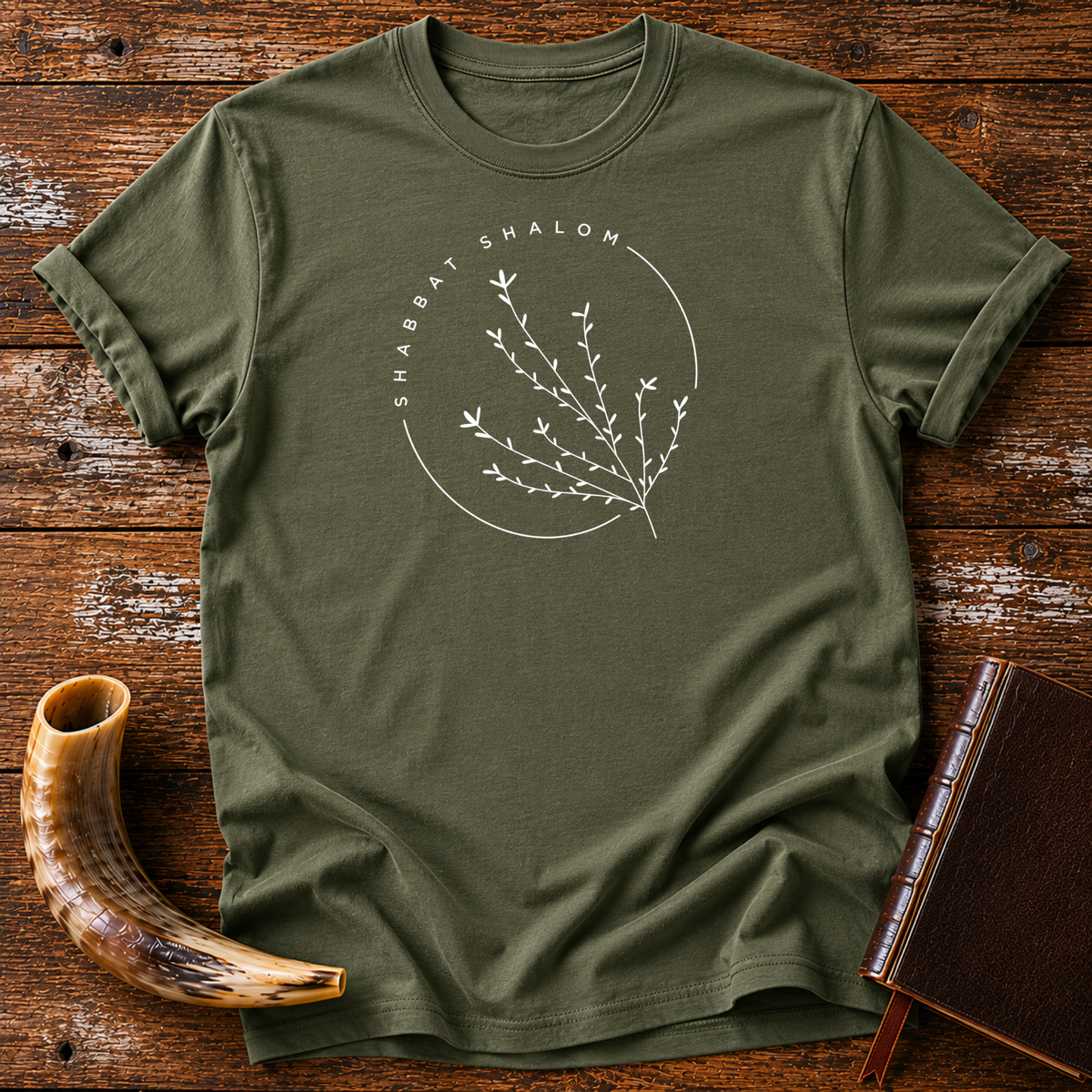 Military green t-shirt with a thin SHABBAT SHALOM circle around a delicate branch, laid flat with a shofar and a leather book
