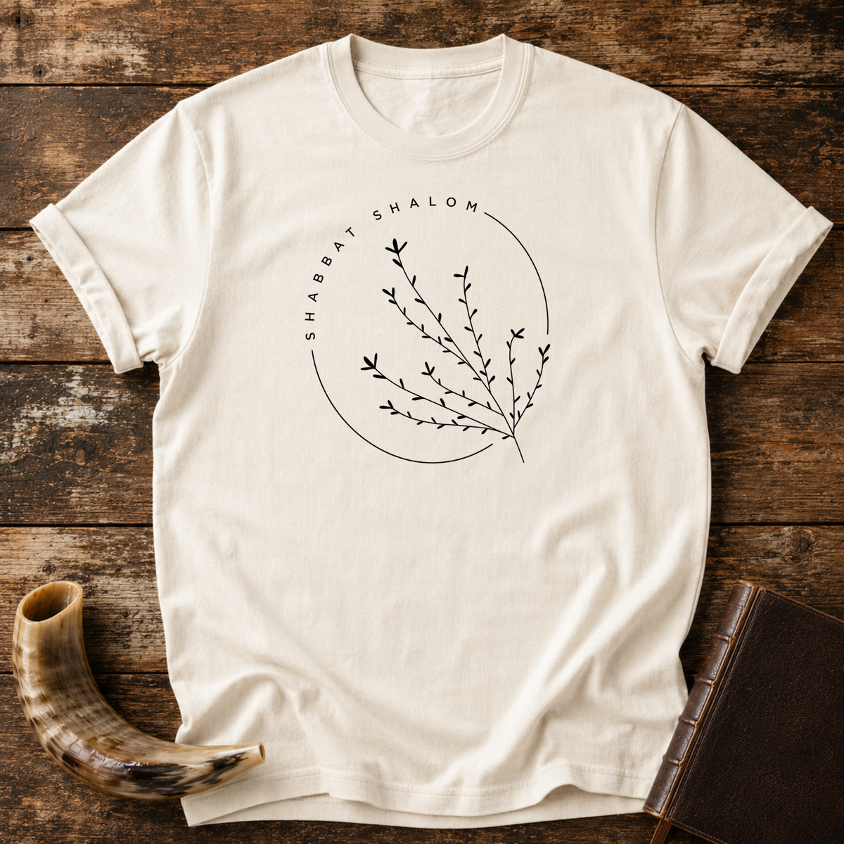 Natural cream t-shirt with a thin SHABBAT SHALOM circle around a delicate branch, laid flat with a shofar and a leather book