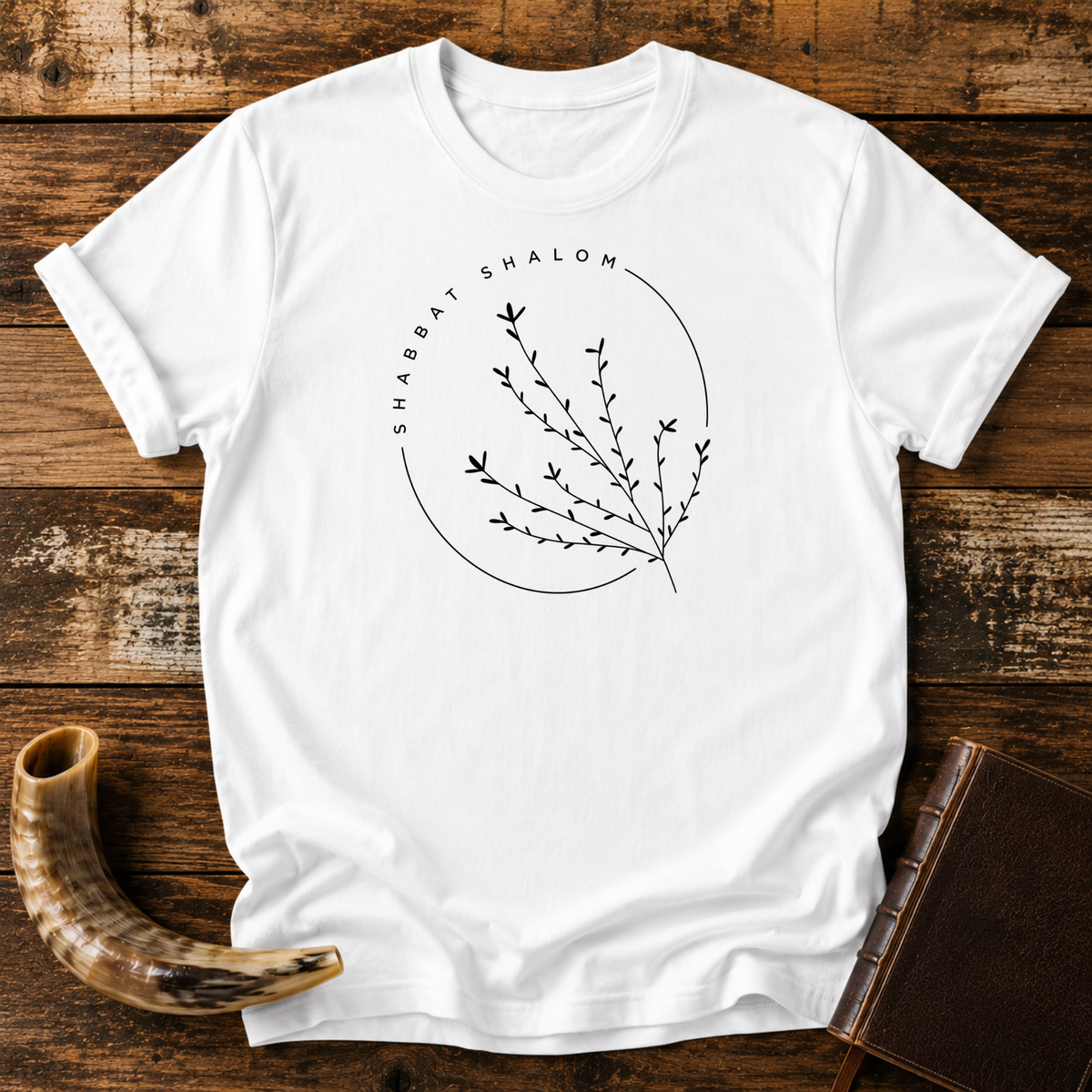 White t-shirt with a thin SHABBAT SHALOM circle around a delicate branch, laid flat with a shofar and a leather book