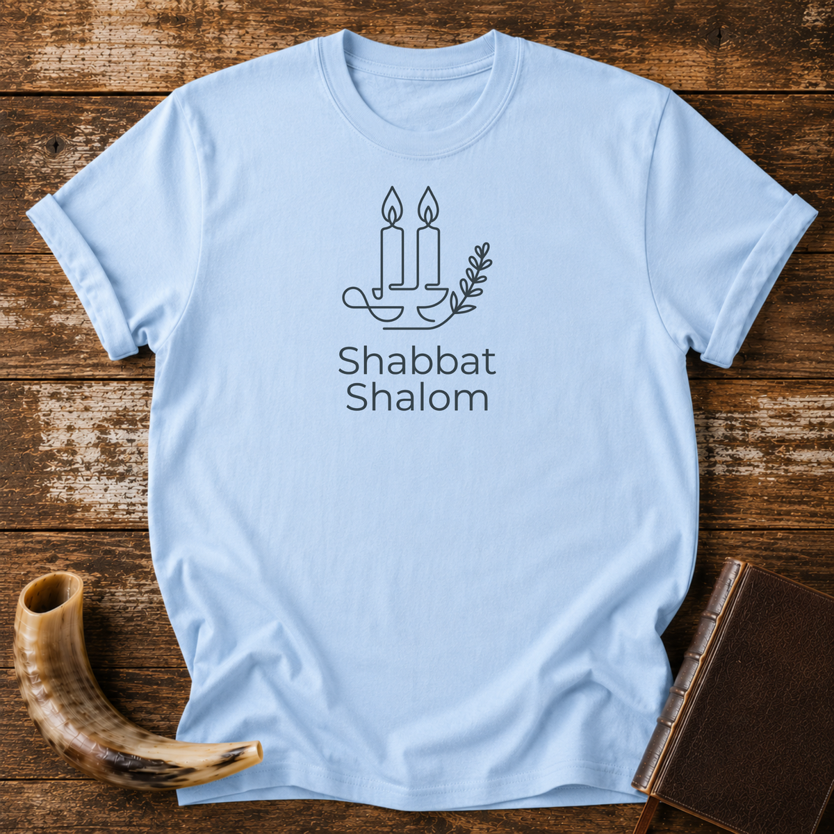Light blue t-shirt with line-art Shabbat candles, an olive branch and Shabbat Shalom text, laid flat with a shofar and a leather book