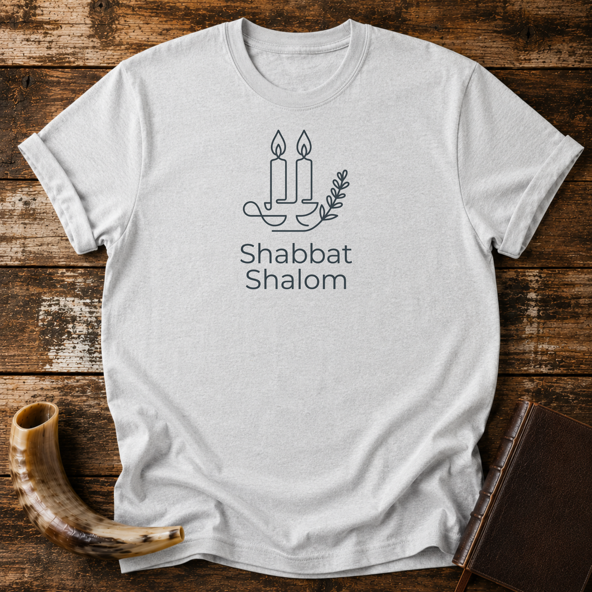 Sport grey t-shirt with line-art Shabbat candles, an olive branch and Shabbat Shalom text, laid flat with a shofar and a leather book