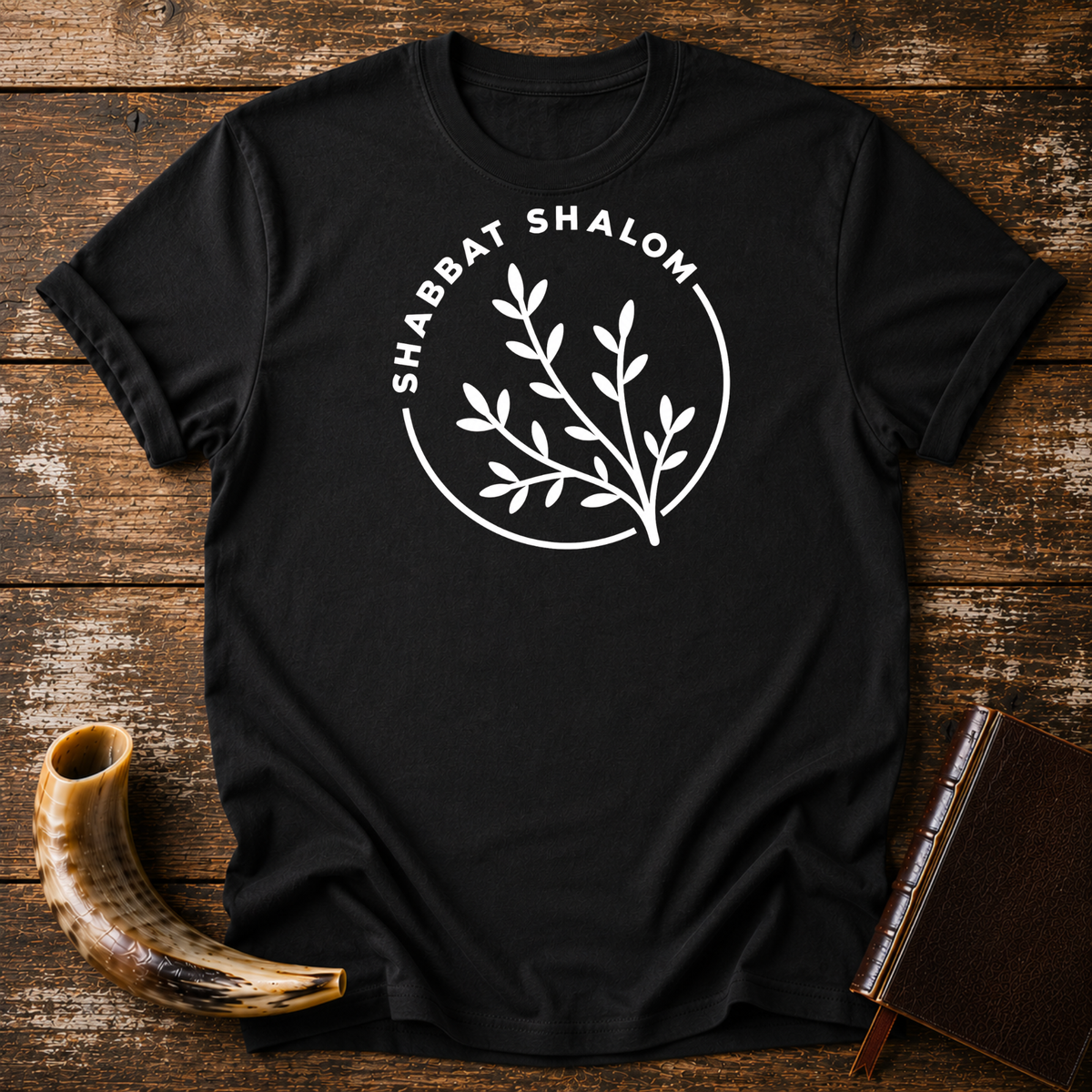 Black t-shirt with a white SHABBAT SHALOM circle around a leafy olive branch, laid flat with a shofar and a leather book