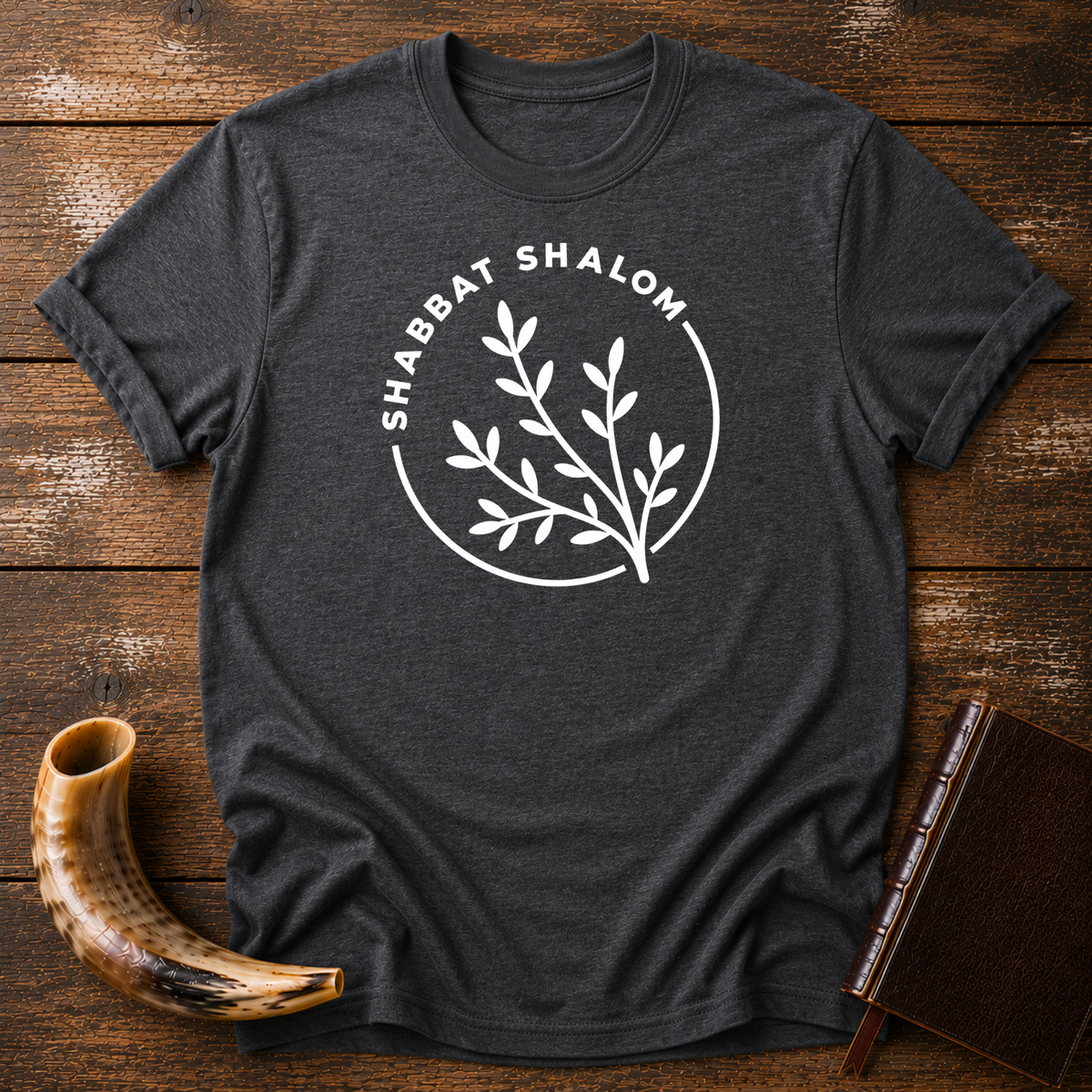 Dark heather gray t-shirt with a white SHABBAT SHALOM circle around a leafy olive branch, laid flat with a shofar and a leather book
