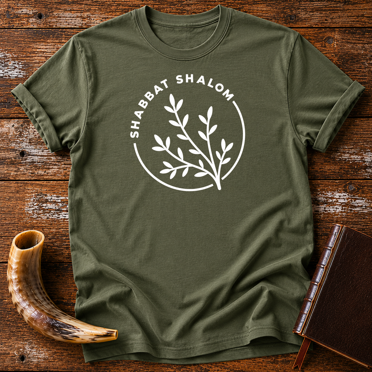 Military green t-shirt with a white SHABBAT SHALOM circle around a leafy olive branch, laid flat with a shofar and a leather book